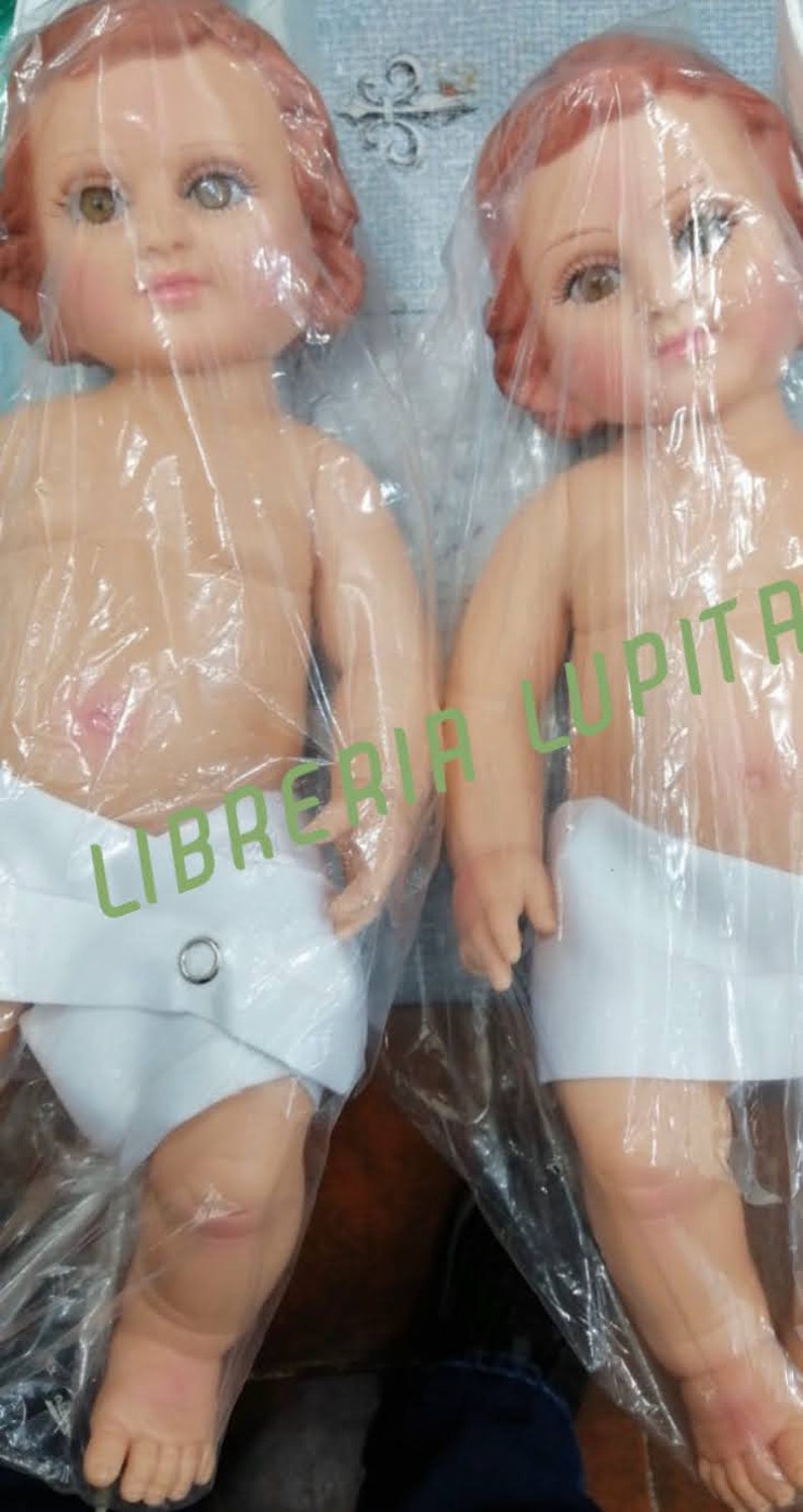 May include: Two baby dolls with painted faces and brown hair, wearing white cloth diapers. The dolls are wrapped in plastic and labeled "Libreria Lupita".