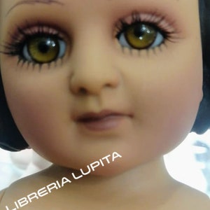 May include: A close-up of a doll's face with brown eyes, painted eyebrows, and a light pink mouth. The doll has a black wig and is wearing a white dress. The text "LIBRERIA LUPITA" is visible in the lower left corner of the image.