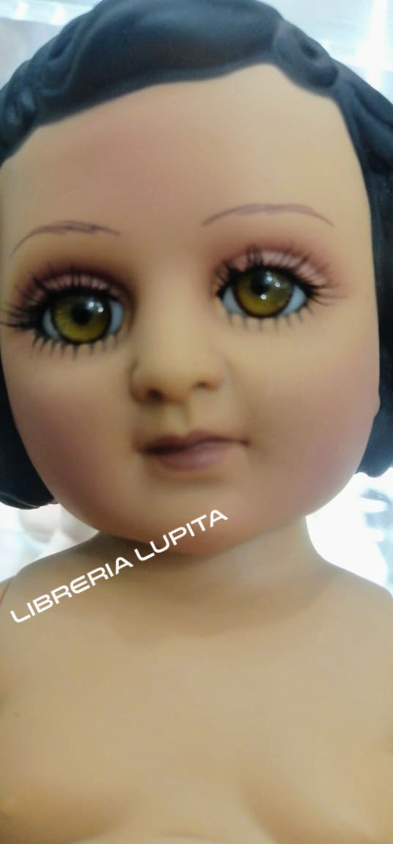 May include: A close-up of a doll's face with brown eyes, painted eyebrows, and a light pink mouth. The doll has a black wig and is wearing a white dress. The text "LIBRERIA LUPITA" is visible in the lower left corner of the image.