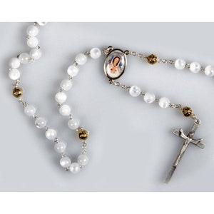 May include: White beaded rosary with silver-tone chain and cross. The rosary features a small oval pendant with a religious image and gold-tone accents. The beads are round and appear to be made of a pearlescent material.