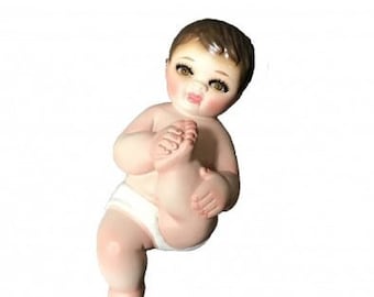 May include: A ceramic figurine of a baby wearing a diaper, sitting with one leg bent and holding their foot. The baby has brown hair, painted eyes, and a rosy complexion.
