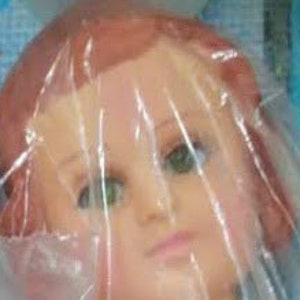 May include: A close-up of a doll's head with brown hair and green eyes. The doll's head is made of plastic and is wrapped in clear plastic.