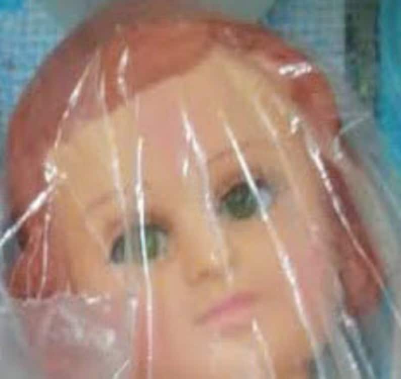 May include: A close-up of a doll's head with brown hair and green eyes. The doll's head is made of plastic and is wrapped in clear plastic.