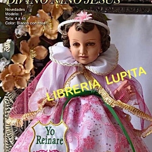 May include: A Niño Dios doll wearing a white and pink lace dress with gold trim, a white ruffled collar, and a pink headband with a flower. The doll features brown hair and holds a shield-shaped patch with the text Yo Reinare in green.
