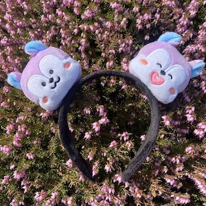 May include: A black headband with two purple and blue plush animal ears. The ears have embroidered faces with pink cheeks and smiling mouths.