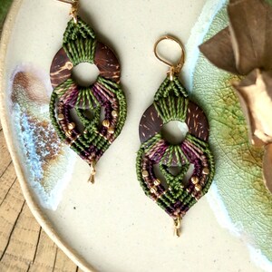 Pode incluir: Um par de brincos de macramé feitos à mão. Cada brinco apresenta uma base de madeira castanha escura com uma abertura circular, adornada com fios verdes e roxos atados. Contas e ganchos dourados completam o design.