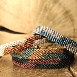 Op de afbeelding: Een stapel handgemaakte macramé-armbanden in verschillende kleuren, waaronder crème, bruin, groen en blauw. De armbanden zijn te zien op een houten oppervlak, met hun ingewikkelde geweven ontwerp. Een stijlvol accessoire.