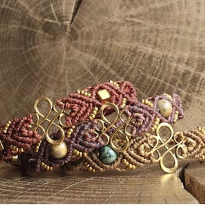 Könnte beinhalten: Drei Makramee-Armbänder aus kunstvoll geknoteten Kordeln in Burgunderrot, Violett und Beige. Jedes Armband enthält kleine goldfarbene Metallperlen, ein zentrales goldfarbenes Drahtelement in Unendlichkeitsform und eine einzelne runde Steinperle.