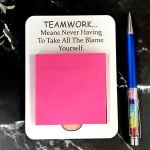 May include: A white rectangular notepad holder with rounded corners featuring the text "TEAMWORK... Means Never Having To Take All The Blame Yourself." A bright pink square sticky note is attached to the centre, paired with a blue ballpoint pen.
