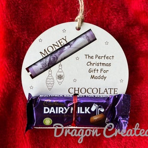 May include: A wooden Christmas bauble with a slot for money and a space for a chocolate bar. The bauble is decorated with stars and the words "MONEY", "The Perfect Christmas Gift For Maddy", "CHOCOLATE", and "Dragon Created".