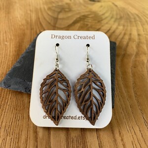 Puede incluir: Un par de pendientes de madera con forma de hoja con intrincados recortes. Los pendientes son marrones y tienen ganchos plateados. Los pendientes se muestran en una tarjeta blanca con el texto "Dragon Created" impreso en ella.