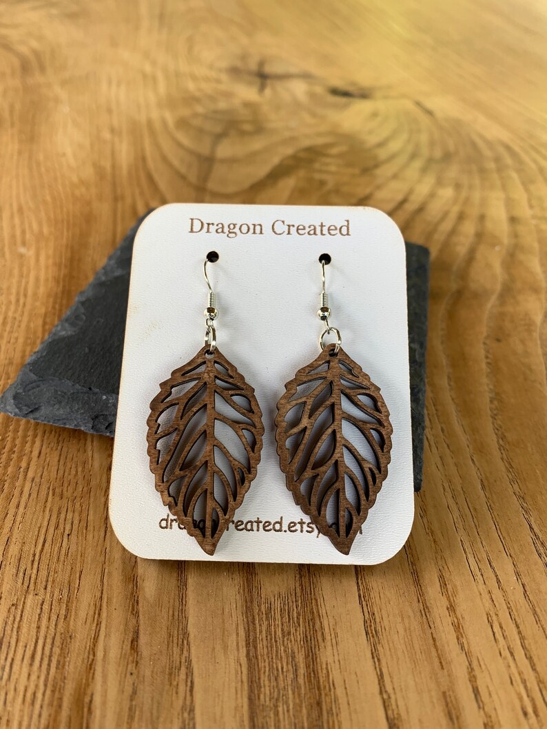 Puede incluir: Un par de pendientes de madera con forma de hoja con intrincados recortes. Los pendientes son marrones y tienen ganchos plateados. Los pendientes se muestran en una tarjeta blanca con el texto "Dragon Created" impreso en ella.
