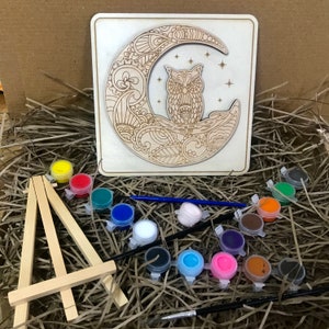May include: A wooden craft kit featuring an owl perched on a crescent moon with intricate details. The kit includes paints, brushes, and a small easel for displaying the finished artwork.
