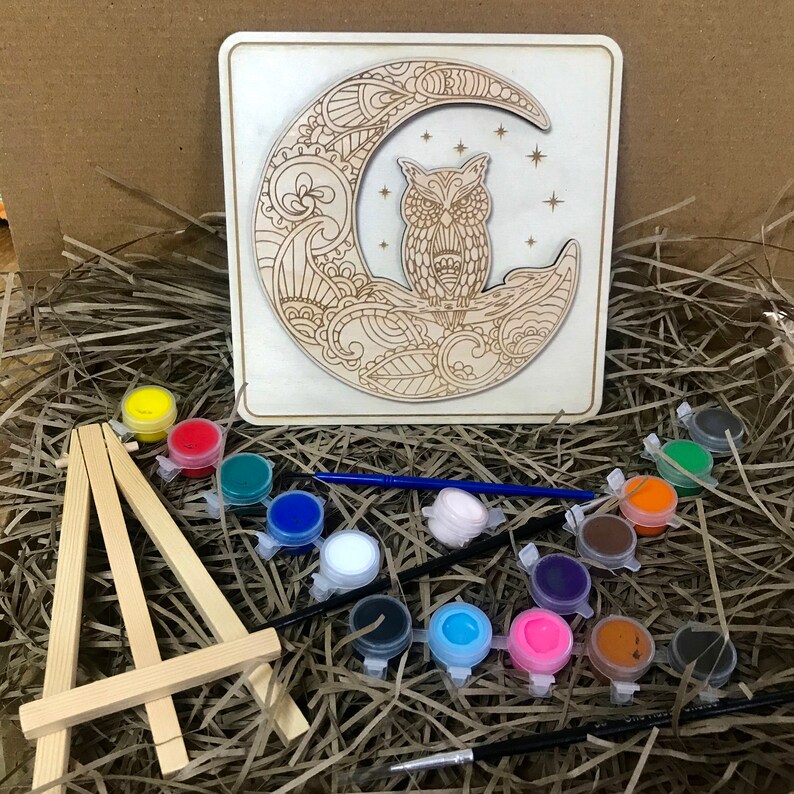 May include: A wooden craft kit featuring an owl perched on a crescent moon with intricate details. The kit includes paints, brushes, and a small easel for displaying the finished artwork.