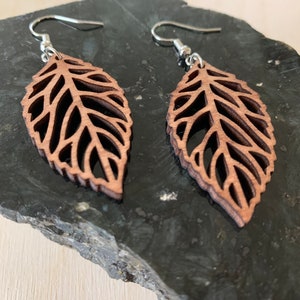 Puede incluir: Un par de pendientes de madera marr&oacute;n en forma de hoja con un delicado dise&ntilde;o de corte. Los pendientes cuelgan de ganchos plateados.