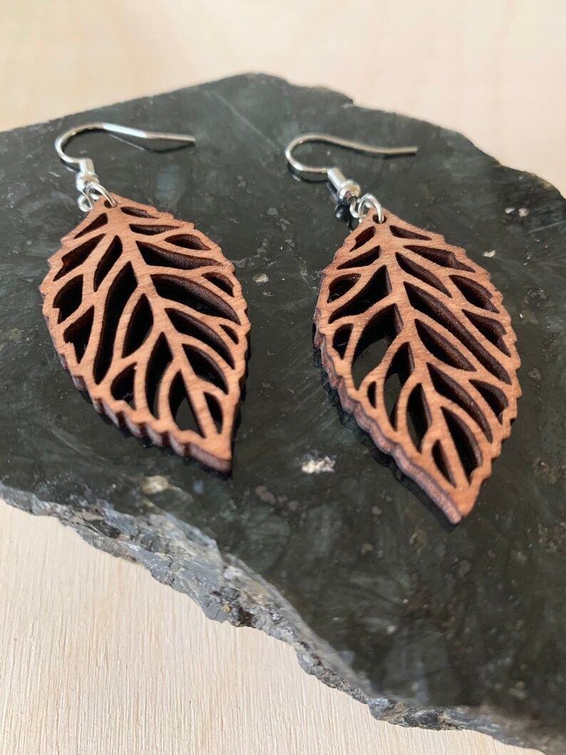 Puede incluir: Un par de pendientes de madera marr&oacute;n en forma de hoja con un delicado dise&ntilde;o de corte. Los pendientes cuelgan de ganchos plateados.