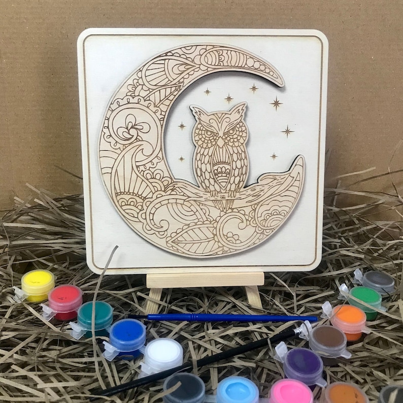 May include: Wooden paint-by-numbers kit featuring an owl perched on a crescent moon with intricate details. The kit includes a wooden easel, paintbrushes, and a variety of acrylic paints in small containers.