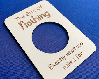 Puede incluir: Una tarjeta de regalo rectangular de madera blanquecina con un gran recorte circular. El texto "The Gift Of Nothing" y "Exactly what you asked for" está grabado en marrón. La tarjeta descansa sobre una superficie azul.