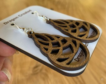 Puede incluir: Un par de pendientes de madera con un diseño geométrico. Los pendientes son marrones y tienen un gancho plateado. Los pendientes están en una tarjeta blanca con el texto "Dragon Created" en ella.