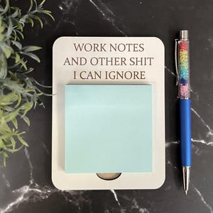 May include: A white notepad with the text "WORK NOTES AND OTHER SHIT I CAN IGNORE" and a light blue sticky note. A blue pen with a rainbow crystal barrel sits to the right. The background is black marble.