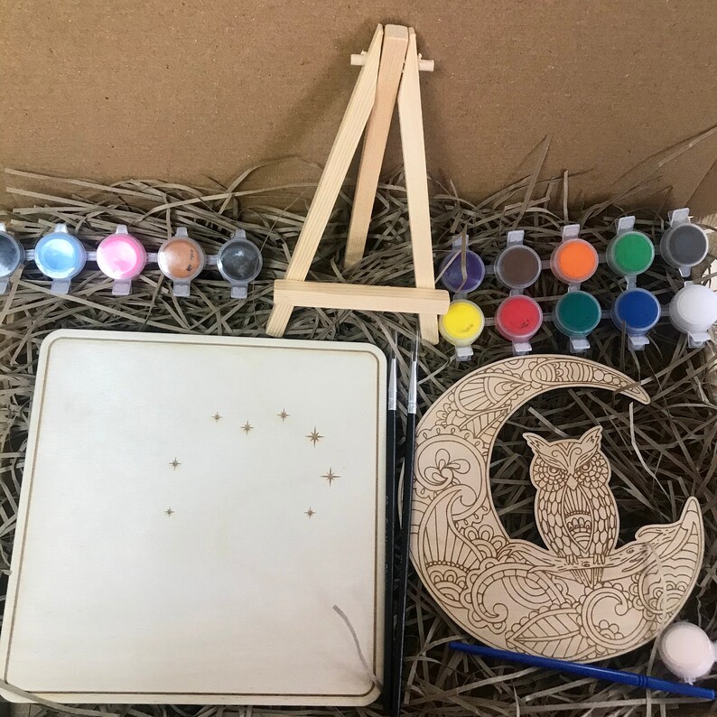 May include: A wooden craft kit featuring a crescent moon with an owl design, a small wooden easel, and a set of paint pots with colorful paint. The kit also includes a blank wooden canvas and a paintbrush.
