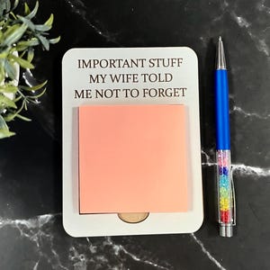 May include: A white rectangular wooden note holder with rounded corners featuring the engraved text IMPORTANT STUFF MY WIFE TOLD ME NOT TO FORGET, holding a square peach sticky note, accompanied by a blue ballpoint pen with a crystal-filled barrel.