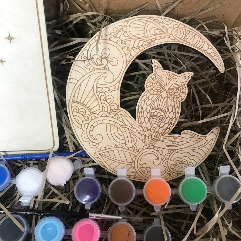 May include: A wooden craft kit featuring an owl perched on a crescent moon with intricate floral designs. The kit includes a variety of paint colors for coloring the design.