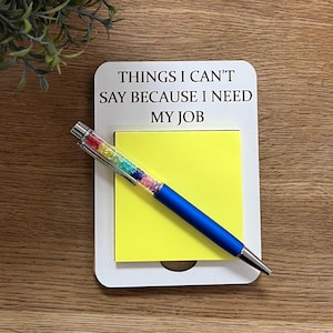 May include: A white rectangular notepad holder with the black text THINGS I CAN'T SAY BECAUSE I NEED MY JOB printed on it. A bright yellow square sticky note is attached to the holder, and a blue ballpoint pen with a rainbow crystal barrel rests on top.