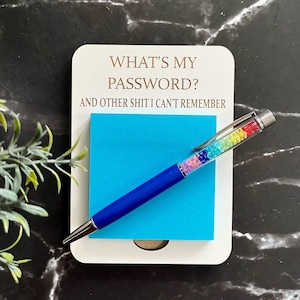 May include: A white rectangular notepad with the text WHAT'S MY PASSWORD? AND OTHER SHIT I CAN'T REMEMBER printed in brown. A bright blue square sticky note is attached to the centre, topped with a blue pen featuring a rainbow crystal-filled barrel.