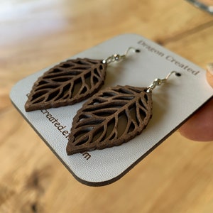 Puede incluir: Un par de pendientes de hoja de madera marr&oacute;n con ganchos plateados. Los pendientes se muestran en una tarjeta blanca con el texto "Dragon Created" impreso en ella.