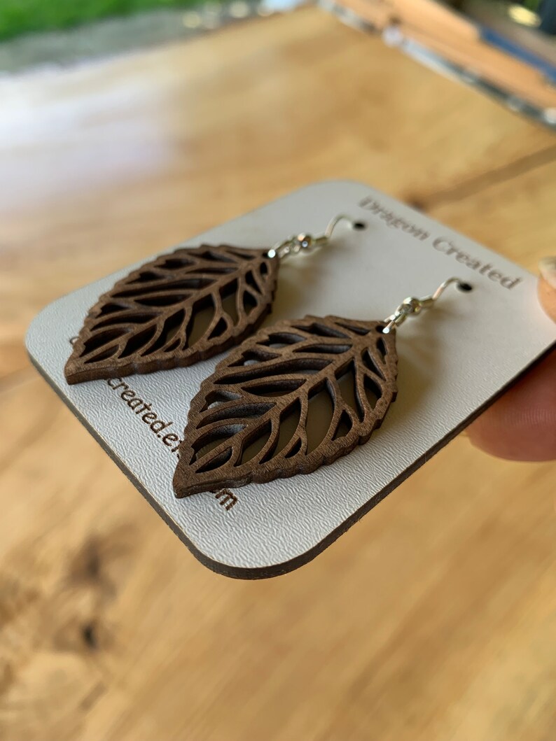 Puede incluir: Un par de pendientes de hoja de madera marr&oacute;n con ganchos plateados. Los pendientes se muestran en una tarjeta blanca con el texto "Dragon Created" impreso en ella.