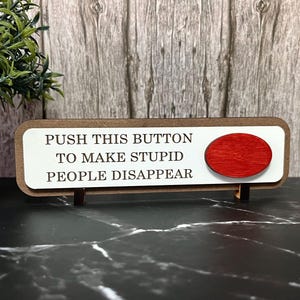 May include: A wooden sign with the text "PUSH THIS BUTTON TO MAKE STUPID PEOPLE DISAPPEAR." The sign has a white background with a brown border and a red button on the right side. The sign is propped up on a black surface.