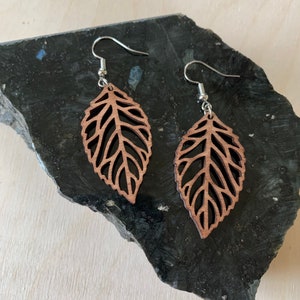 May include: A pair of brown wooden leaf earrings with a delicate cut-out design. The earrings are suspended from silver hooks.