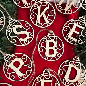 May include: A set of wooden Christmas decorations featuring a decorative swirl design and a letter cut out in the centre. The ornaments are in natural wood colours and are hanging from a red background.