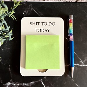 May include: A cream notepad with the text "SHIT TO DO TODAY" and a bright green sticky note. A blue pen with a rainbow crystal barrel sits beside the notepad on a black marble surface.