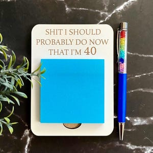 May include: A white rectangular note holder with the engraved text SHIT I SHOULD PROBABLY DO NOW THAT I'M 40, featuring a bright blue square sticky note and a blue ballpoint pen with a clear barrel filled with small, colourful rainbow crystals.