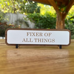May include: A wooden desk sign with the text "FIXER OF ALL THINGS" in a brown font. The sign has a white centre and a brown border. It is supported by two small black stands.