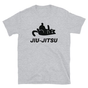 Puede incluir: Una camiseta gris con una ilustración negra de un gato haciendo un movimiento de jiu-jitsu. El texto "JIU-JITSU" está impreso en negro debajo del gato.