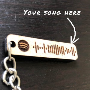 Peut inclure: Un porte-clés en bois avec une chaîne argentée et un code-barres Spotify gravé dessus. Le texte "YOUR SONG HERE" est écrit au-dessus du code-barres.