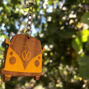 Peut inclure: Un porte-clés en bois en forme de Volkswagen jaune avec un contour brun. Le porte-clés est suspendu à une chaîne argentée.