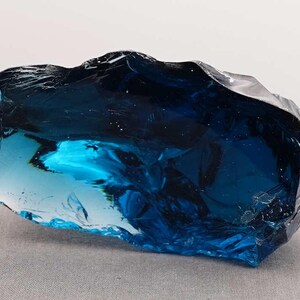 Puede incluir: Un gran trozo de vidrio azul de forma irregular. El vidrio tiene una cualidad transl&uacute;cida, con &aacute;reas de azul marino profundo y turquesa m&aacute;s claro. La superficie tiene un aspecto rugoso y texturizado, que capta la luz y crea reflejos. La forma general es un trozo s&oacute;lido y sustancial.