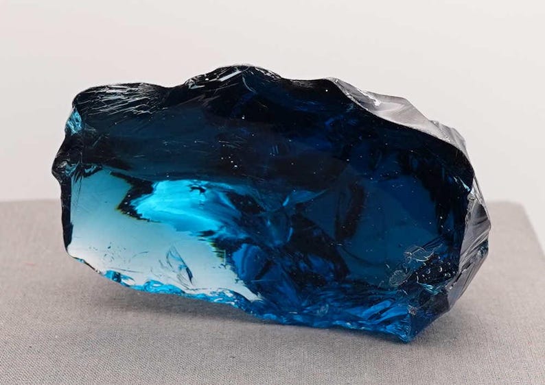 Puede incluir: Un gran trozo de vidrio azul de forma irregular. El vidrio tiene una cualidad transl&uacute;cida, con &aacute;reas de azul marino profundo y turquesa m&aacute;s claro. La superficie tiene un aspecto rugoso y texturizado, que capta la luz y crea reflejos. La forma general es un trozo s&oacute;lido y sustancial.