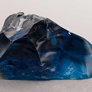 Puede incluir: Un gran trozo de vidrio azul de forma irregular. El vidrio tiene una superficie brillante y reflectante con diferentes tonos de azul y negro. El objeto est&aacute; sobre un fondo neutro, resaltando su color y forma.
