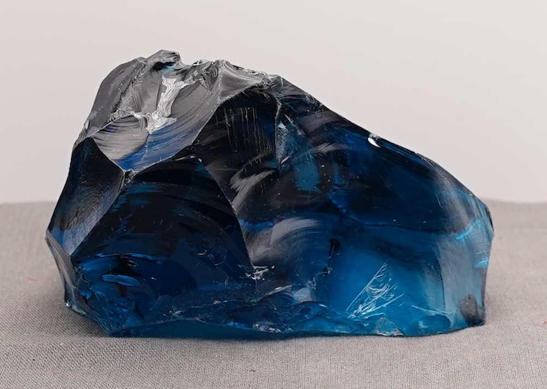 Puede incluir: Un gran trozo de vidrio azul de forma irregular. El vidrio tiene una superficie brillante y reflectante con diferentes tonos de azul y negro. El objeto est&aacute; sobre un fondo neutro, resaltando su color y forma.