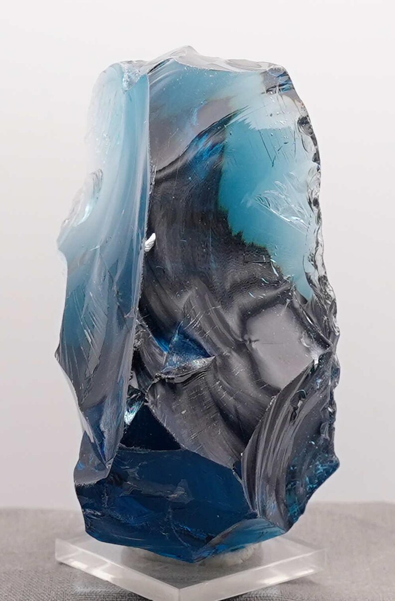 Puede incluir: Escultura abstracta de vidrio en tonos azules y grises. El vidrio es transl&uacute;cido y tiene una superficie irregular y texturizada. Montada sobre una base de acr&iacute;lico transparente.