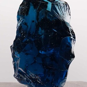 Puede incluir: Una pieza grande de vidrio azul oscuro de forma irregular, posiblemente un objeto decorativo o una obra de arte. El vidrio tiene una superficie texturizada con fracturas internas, montado sobre una base de acr&iacute;lico transparente. El color es un azul rico y saturado.