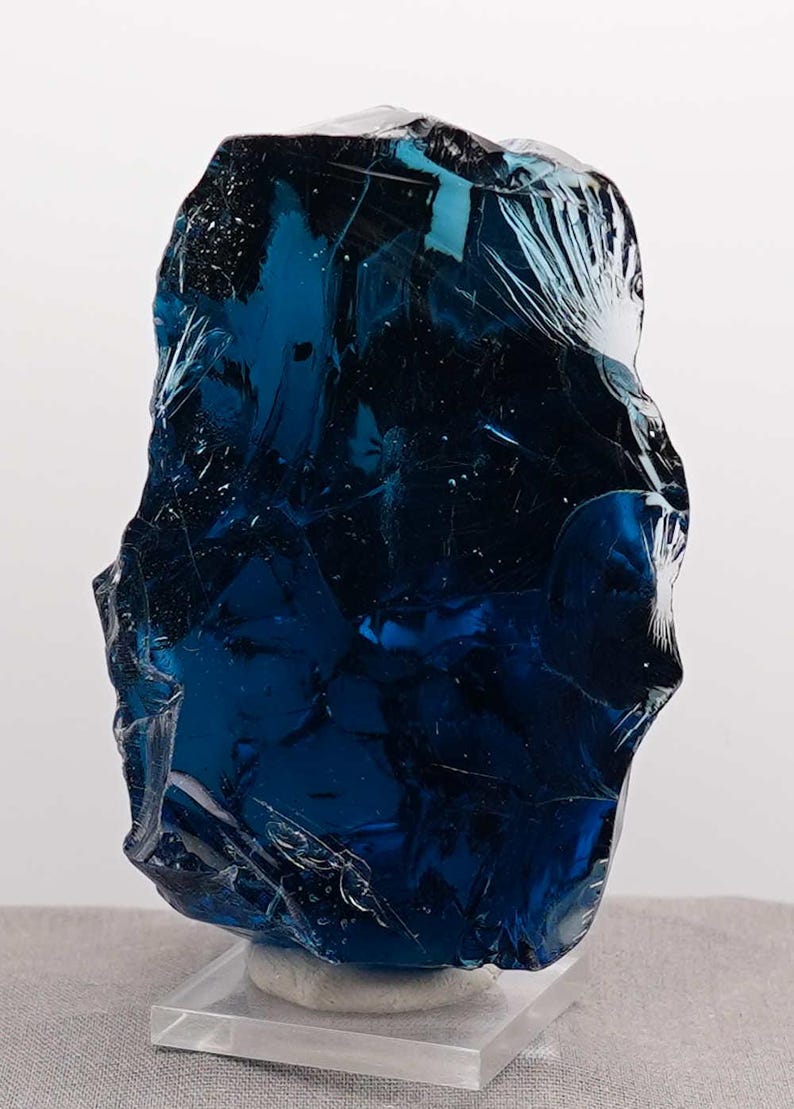 Puede incluir: Una pieza grande de vidrio azul oscuro de forma irregular, posiblemente un objeto decorativo o una obra de arte. El vidrio tiene una superficie texturizada con fracturas internas, montado sobre una base de acr&iacute;lico transparente. El color es un azul rico y saturado.