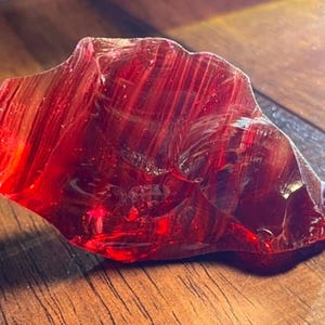 May include: A striking, translucent red glass rock with an uneven, natural form. The polished surface catches the light, revealing internal patterns. The rock rests on a wooden surface, emphasizing its colour and texture. A decorative item.