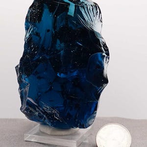 Puede incluir: Una pieza grande de vidrio azul oscuro, de forma irregular, posiblemente escoria, se exhibe sobre un soporte de acr&iacute;lico transparente. Una moneda est&aacute; al lado del vidrio para comparar el tama&ntilde;o. El vidrio tiene una superficie pulida y reflectante.