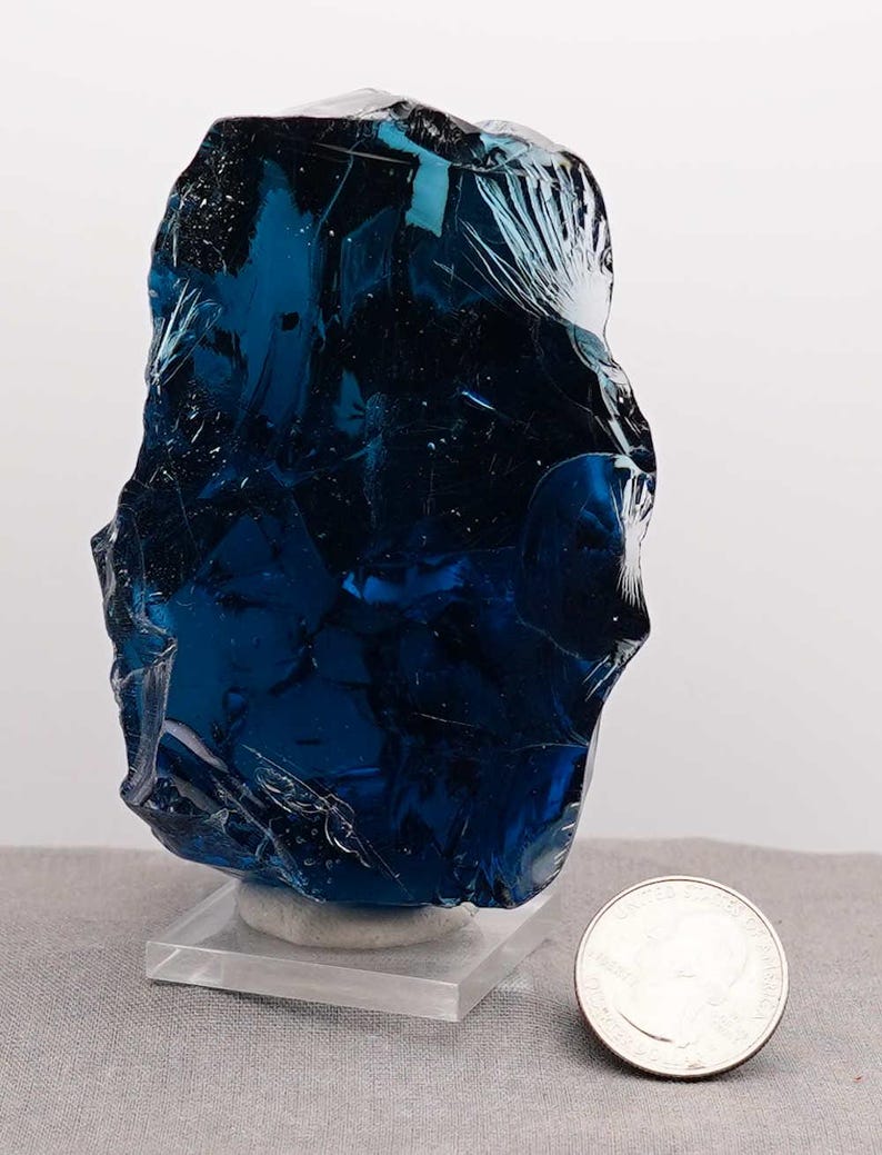 Puede incluir: Una pieza grande de vidrio azul oscuro, de forma irregular, posiblemente escoria, se exhibe sobre un soporte de acr&iacute;lico transparente. Una moneda est&aacute; al lado del vidrio para comparar el tama&ntilde;o. El vidrio tiene una superficie pulida y reflectante.